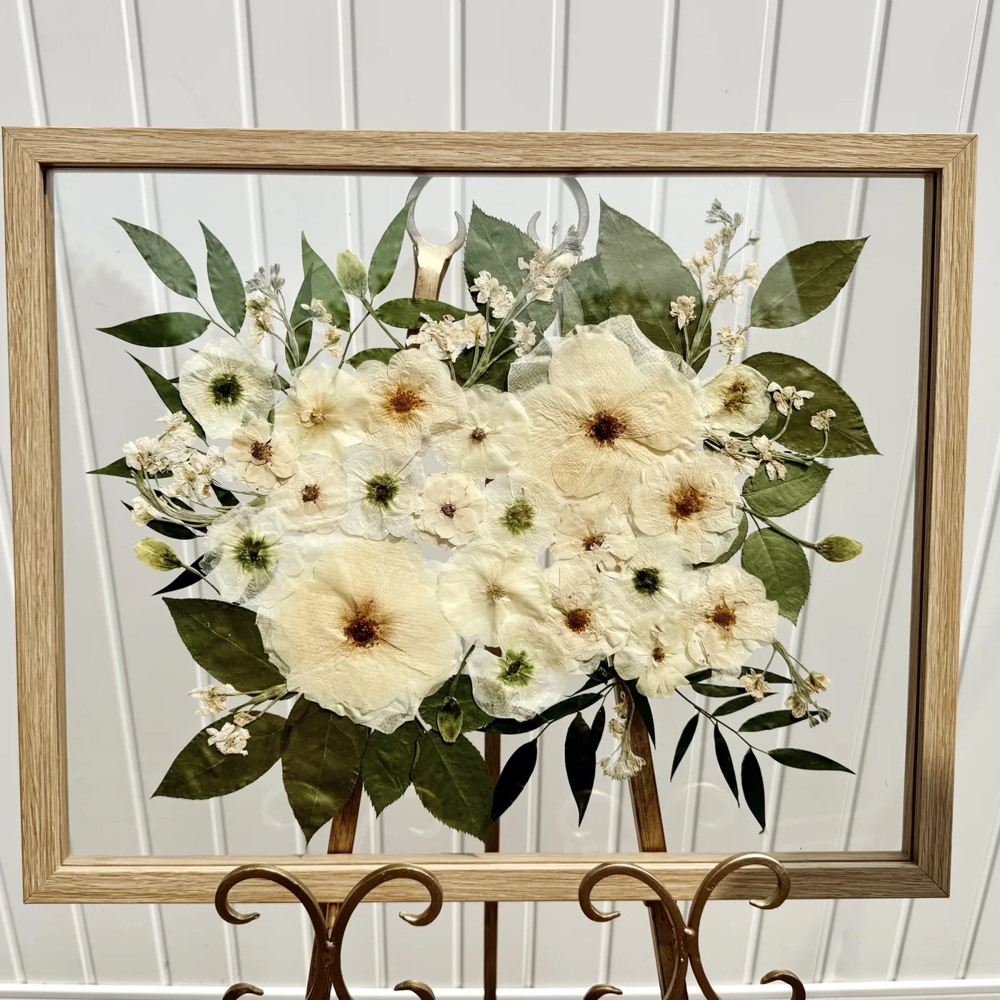 A wood frame of cream pressed flowers and greenery on an easel